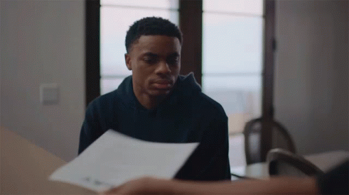 A Man In A Black Hoodie Is Sitting At A Table With A Piece Of Paper In Front Of Him GIF
