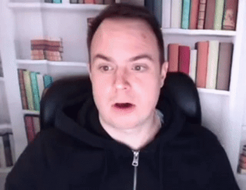 A Man In A Black Hoodie Is Sitting In Front Of A Bookshelf And Making A Funny Face . GIF