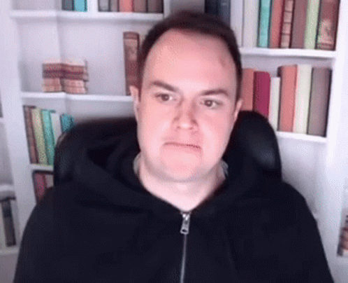 A Man In A Black Hoodie Is Sitting In Front Of A Bookshelf With Books . GIF