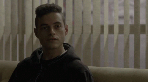 A Man In A Black Hoodie Is Sitting On A Couch In Front Of Vertical Blinds GIF