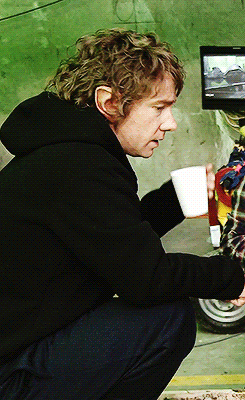 A Man In A Black Hoodie Is Squatting Down Holding A Cup Of Coffee GIF