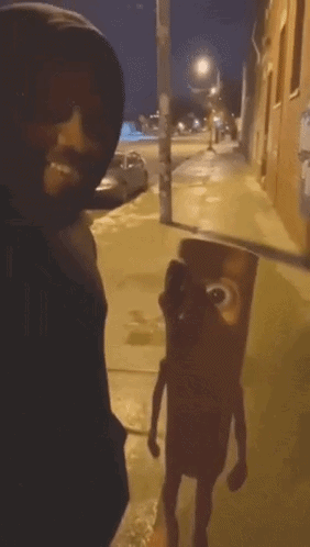 A Man In A Black Hoodie Is Standing Next To A Cartoon Character On The Sidewalk GIF