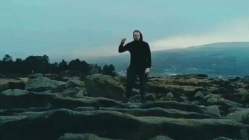 A Man In A Black Hoodie Is Standing On A Rocky Cliff With A Mountain In The Background . GIF