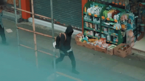 A Man In A Black Hoodie Is Walking Down A Sidewalk In Front Of A Store . GIF