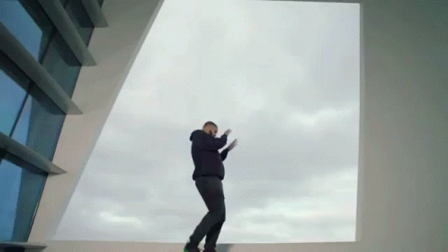A Man In A Black Hoodie Stands In Front Of A Large Window GIF