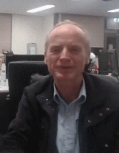 A Man In A Black Jacket And Blue Shirt Is Sitting At A Desk In An Office GIF