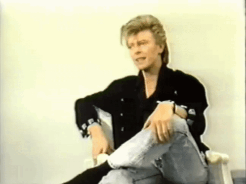 A Man In A Black Jacket And Jeans Is Sitting In A Chair With His Legs Crossed . GIF