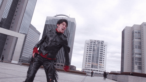 A Man In A Black Jacket And Red Gloves Stands In Front Of A City Skyline GIF