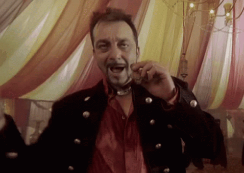 A Man In A Black Jacket And Red Shirt Is Holding Something In His Mouth While Standing In Front Of A Tent GIF