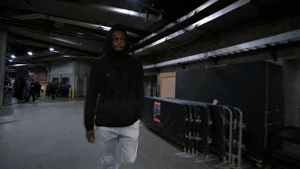 A Man In A Black Jacket And White Pants Is Walking Through A Dark Room GIF