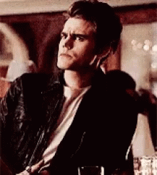 A Man In A Black Jacket And White Shirt Is Sitting In A Room . GIF
