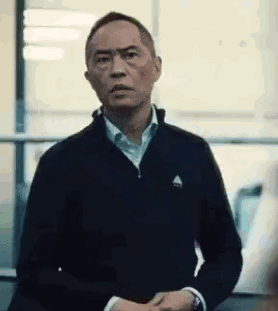 A Man In A Black Jacket And White Shirt Is Standing In Front Of A Window And Making A Funny Face . GIF