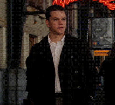 A Man In A Black Jacket And White Shirt Is Walking Down A Street In Front Of A Sign That Says ' Coca Cola ' GIF