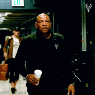 A Man In A Black Jacket Holds A Cup Of Coffee In Front Of An Exit Sign GIF