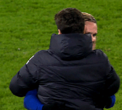 A Man In A Black Jacket Is Hugging Another Man In A Blue Shirt GIF