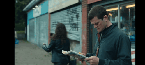 a man in a black jacket is reading a newspaper while a woman looks on