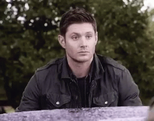 A Man In A Black Jacket Is Sitting At A Table In A Park And Looking At The Camera . GIF
