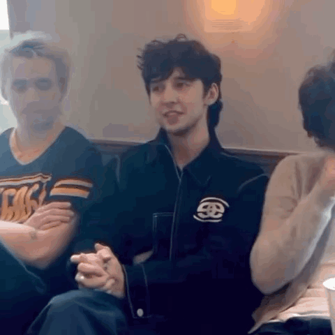 A Man In A Black Jacket Is Sitting In A Chair With His Hands Folded And Talking To A Group Of People . GIF