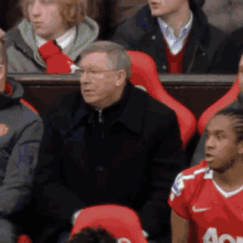 A Man In A Black Jacket Is Sitting In A Stadium With Other People GIF