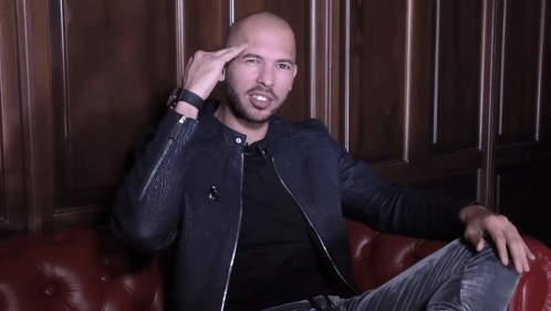 A Man In A Black Jacket Is Sitting On A Red Leather Couch With His Arms Outstretched GIF
