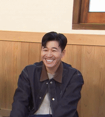 A Man In A Black Jacket Is Smiling In Front Of A Wooden Wall . GIF