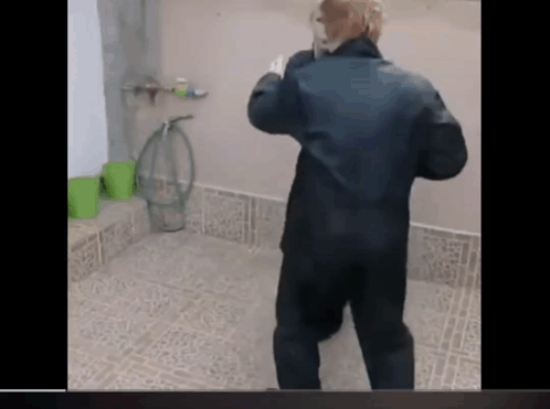 a man in a black jacket is standing in a bathroom talking on a phone .