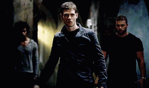 A Man In A Black Jacket Is Standing In A Dark Hallway With Other Men GIF