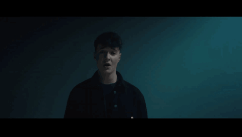 A Man In A Black Jacket Is Standing In A Dark Room Looking At The Camera GIF