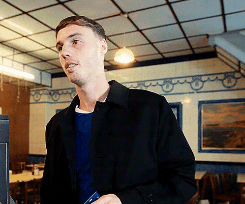 A Man In A Black Jacket Is Standing In A Room With A Picture On The Wall GIF