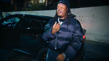 A Man In A Black Jacket Is Standing In Front Of A Car . GIF