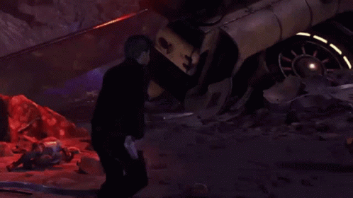 A Man In A Black Jacket Is Standing In Front Of A Destroyed Car GIF
