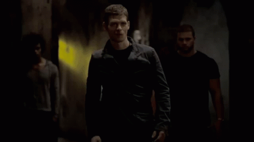 A Man In A Black Jacket Is Walking Down A Dark Hallway With Two Other Men . GIF