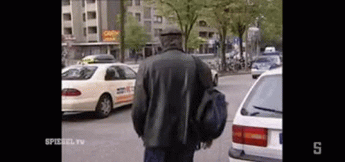 A Man In A Black Jacket Is Walking Down A Street Next To A Taxi . GIF