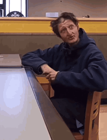 A Man In A Black Jacket Sits At A Table With His Arms Crossed GIF