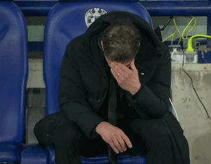 A Man In A Black Jacket Sits In A Blue Chair With His Head In His Hands GIF