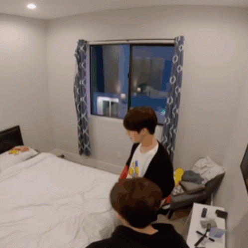 A Man In A Black Jacket Stands In A Bedroom Next To A Bed GIF