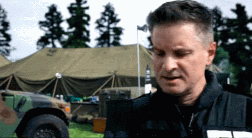 A Man In A Black Jacket Stands In Front Of A Military Vehicle With The Word Army On It GIF