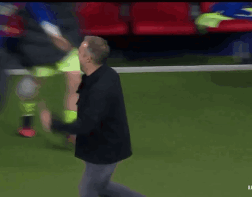 A Man In A Black Jacket Stands On A Soccer Field Surrounded By Players GIF