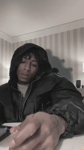 A Man In A Black Jacket With A Hood Is Sitting At A Table GIF