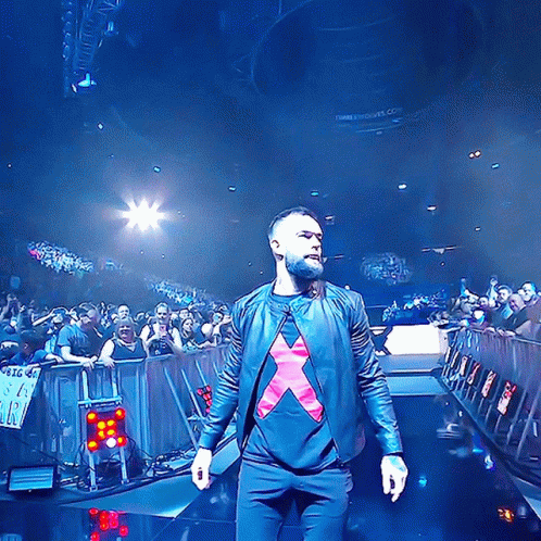 A Man In A Black Jacket With A Red X On His Shirt Stands On A Stage GIF
