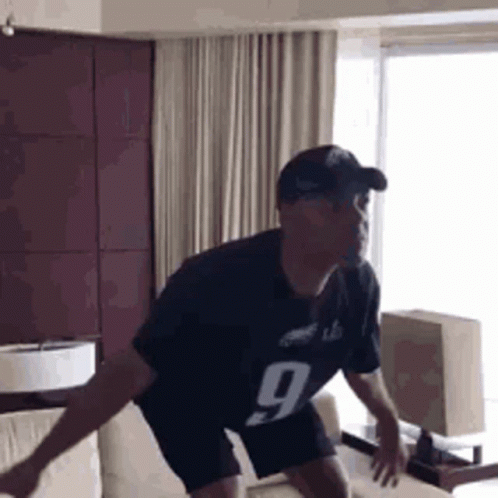A Man In A Black Jersey With The Number 9 On It Is Jumping On A Couch In A Living Room . GIF