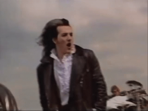 A Man In A Black Leather Jacket And White Shirt Is Standing In Front Of A Motorcycle . GIF