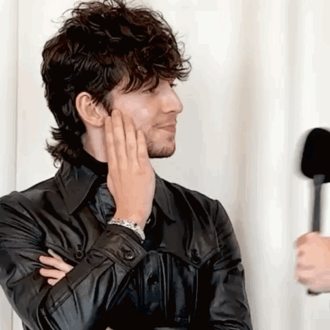 A Man In A Black Leather Jacket Is Holding His Hand To His Face While Talking Into A Microphone . GIF