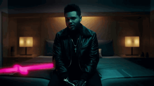 A Man In A Black Leather Jacket Is Sitting On A Bed With A Pink Light Behind Him GIF