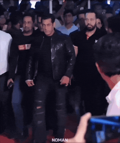 A Man In A Black Leather Jacket Is Walking On A Red Carpet With The Word Noman On The Bottom Right GIF