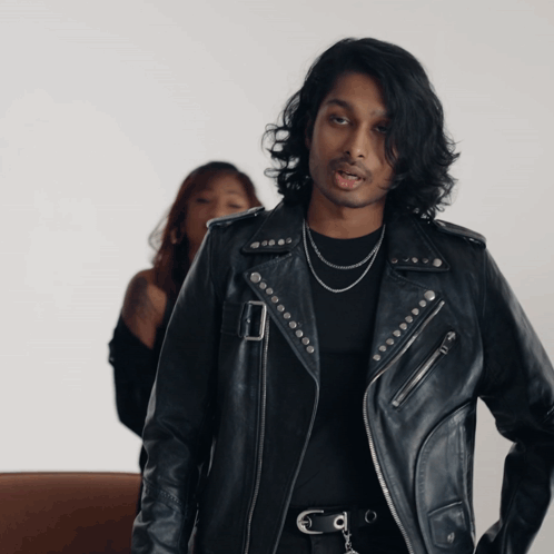 A Man In A Black Leather Jacket Stands Next To A Woman GIF