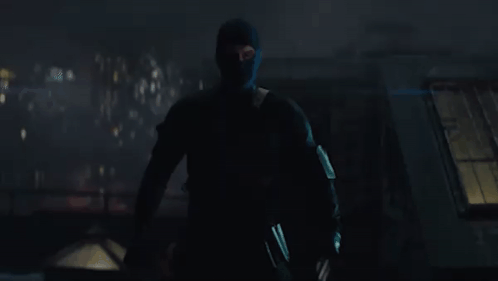 A Man In A Black Mask Is Standing In The Dark Holding A Gun . GIF