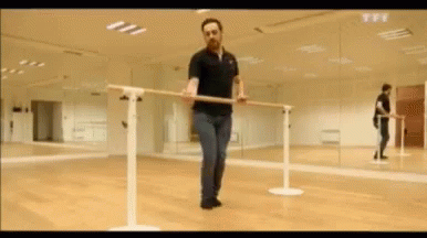 A Man In A Black Polo Shirt Is Holding A Bar In A Dance Studio . GIF