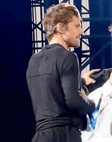 A Man In A Black Shirt And Black Shorts Is Standing On A Stage With A Microphone On His Head . GIF