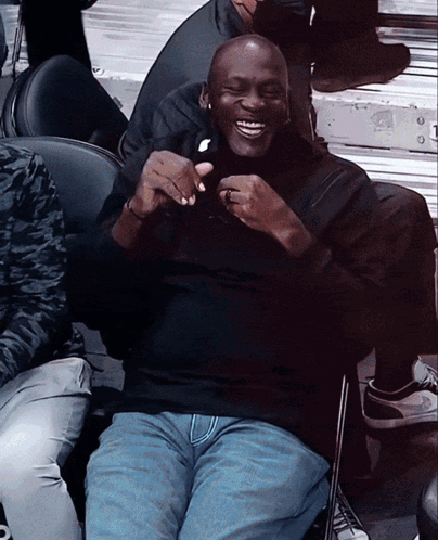 A Man In A Black Shirt And Blue Jeans Sits In A Chair And Smiles GIF